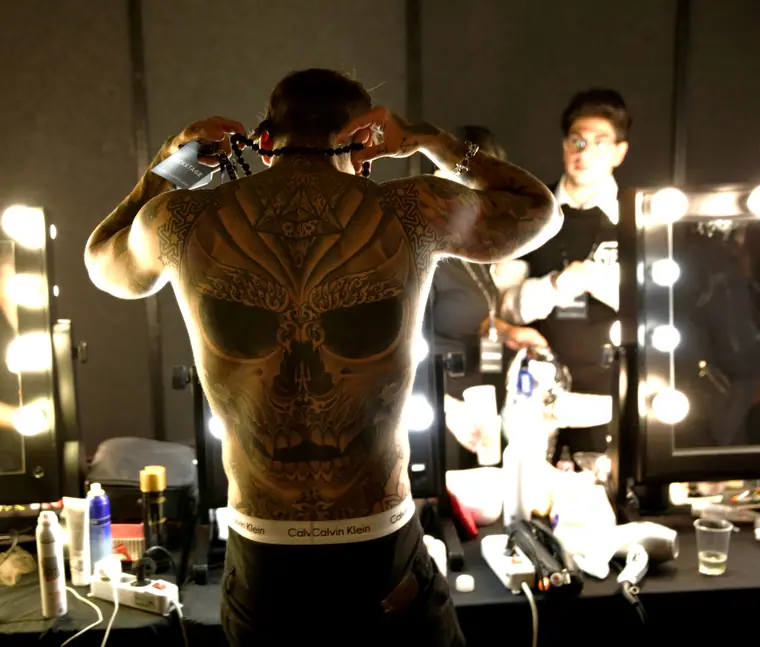 A tattooed back reflected in a bulb-lit backstage mirror
